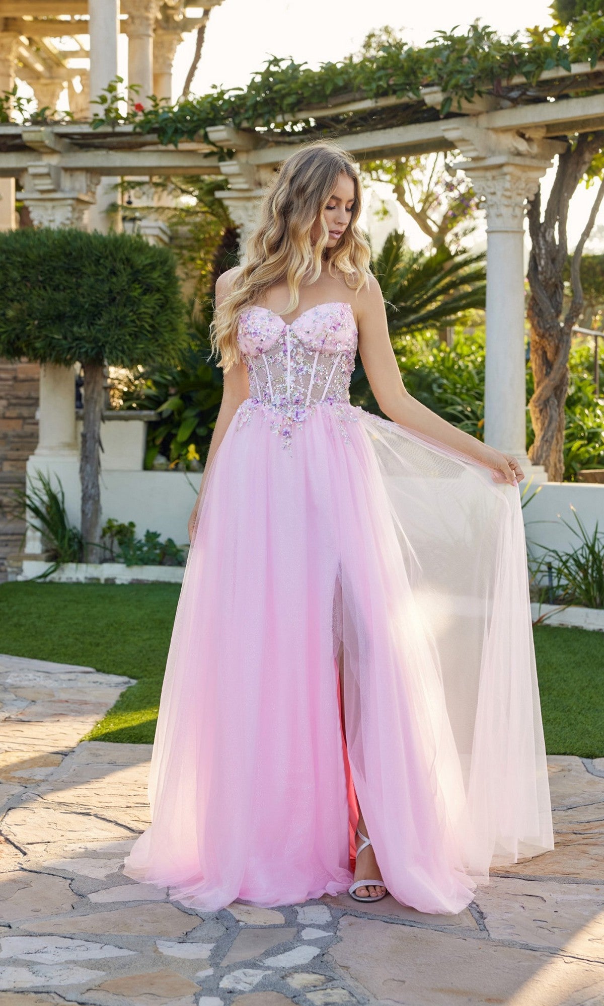 A woman in the Long Prom Dress: Juliet JT2682A in baby pink, featuring a strapless glitter bodice with beads and a tulle skirt, stands outdoors on a stone path, holding one side of her gown.
